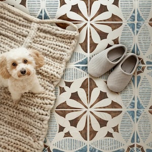 May include: A small, light-colored dog with floppy ears sits on a chunky, cream-colored knit blanket. Next to the blanket, a pair of light gray, textured slip-on slippers rests on a floor featuring a repeating geometric tile pattern in brown, blue, and white.