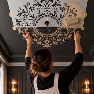 May include: A large white stencil with a black intricate floral and scrollwork pattern taped to a dark ceiling. A paint roller applies a light beige paint through the stencil onto the black surface, revealing a decorative medallion design.