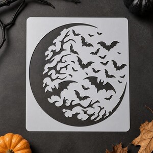 May include: A square stencil featuring a large crescent moon shape filled with cutouts of numerous flying bats and swirling cloud patterns. The design is rendered in a light gray material with dark negative space showing through the cutouts.