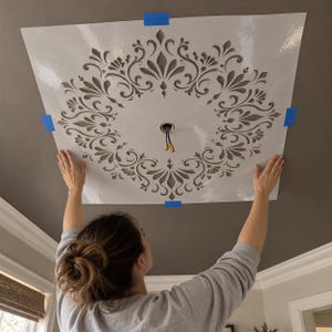 May include: A square white stencil with a symmetrical, intricate floral and scrollwork pattern featuring a central circular cutout. The stencil is secured to a dark gray ceiling with blue painter's tape, positioned around a light fixture opening.