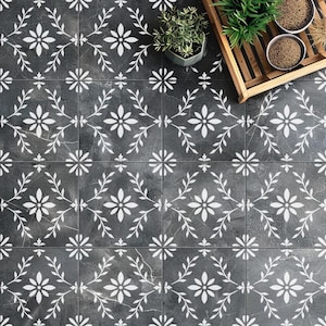 May include: Overhead view of a tiled floor with a repeating floral pattern. The tiles are dark gray with white floral and leaf designs. A wooden tray with small potted plants and bowls sits in the upper right corner, adding a touch of greenery to the scene.
