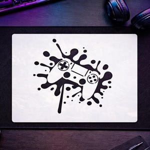 May include: A rectangular white mouse pad featuring a black graphic print of a video game controller centered within a black ink splatter design. The controller illustration includes standard directional buttons, analog sticks, and action buttons.