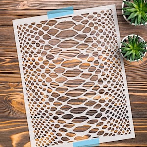 May include: A rectangular white stencil sheet featuring a repeating snake skin pattern with elongated, diamond-shaped cutouts. The stencil is secured at the top and bottom edges with strips of light blue painter's tape on a dark wooden surface.