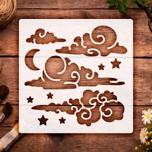 May include: A white stencil featuring a celestial design with clouds, stars, a crescent moon, and a sun. The stencil is placed on a wooden surface, surrounded by paintbrushes, paint, and other craft supplies. The design is ideal for DIY projects.