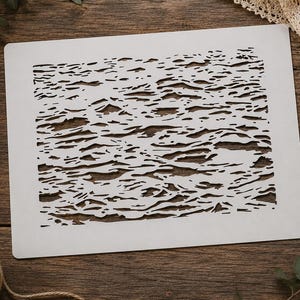 May include: A rectangular white stencil featuring a complex, irregular cut-out pattern of horizontal, wavy, and fragmented shapes that resemble the texture of rippling water or abstract organic layers across the entire surface of the sheet.
