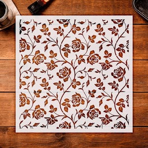 May include: A white square stencil featuring a repeating pattern of roses and leaves in a brown color. The floral design is cut out, creating a negative space effect. The stencil is placed on a wooden surface.