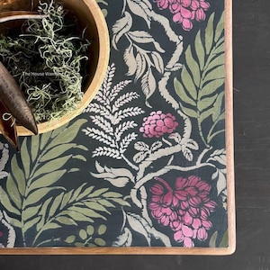 a dark green table with a floral pattern in shades of pink and green. A wooden bowl filled with moss and a pair of scissors are placed on the table.