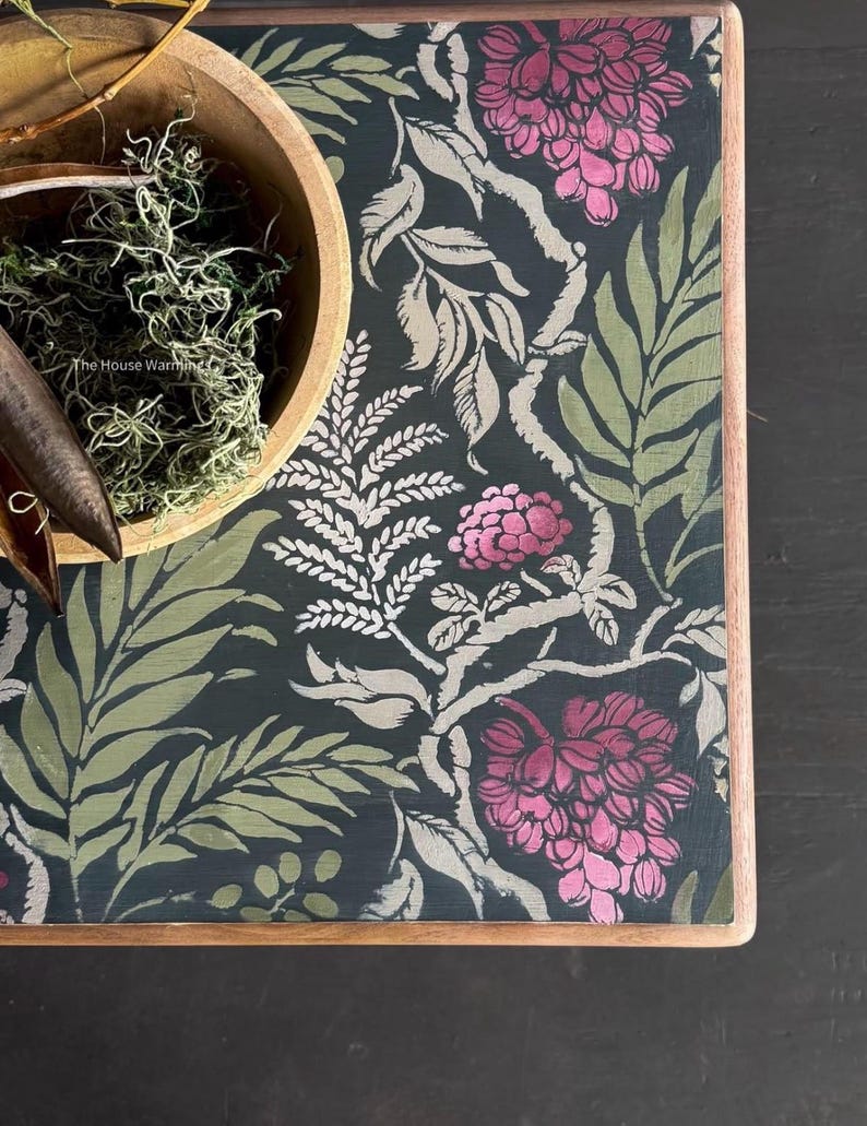 a dark green table with a floral pattern in shades of pink and green. A wooden bowl filled with moss and a pair of scissors are placed on the table.