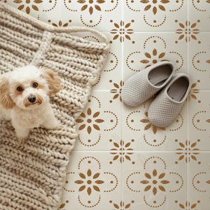 May include: A small cream-colored dog with floppy ears sits on a thick, chunky knit beige blanket next to a pair of light gray textured slip-on slippers. The items are placed on a white tiled floor featuring a repeating brown floral pattern.