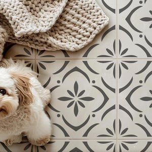 May include: A small, light-colored, fluffy dog sits on a floor covered in square tiles featuring a repeating dark gray floral pattern on a cream background. A thick, beige, chunky-knit blanket is draped across the upper left corner of the tiles.