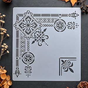 May include: A square, semi-transparent stencil sheet featuring intricate corner designs. The pattern includes a large, ornate geometric border with floral motifs, circular medallions, and a smaller, separate fleur-de-lis corner accent.