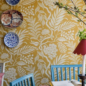 May include: Mustard yellow wallpaper with a white botanical pattern featuring leaves and pinecones. Three decorative plates are mounted on the wall. Two lamps with scalloped and polka-dot shades sit on a table with blue chairs and a red plaid pillow.