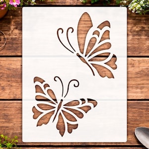 May include: A rectangular white stencil featuring two die-cut butterfly designs. The top butterfly is angled upward, while the bottom butterfly is positioned facing forward with symmetrical wings. Both butterflies display intricate cut-out patterns.