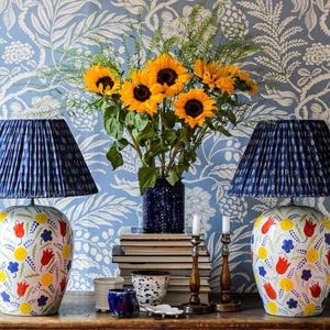a table with two blue lamps, a vase of yellow sunflowers, and a stack of books. The table is set against a blue and white floral wallpaper.