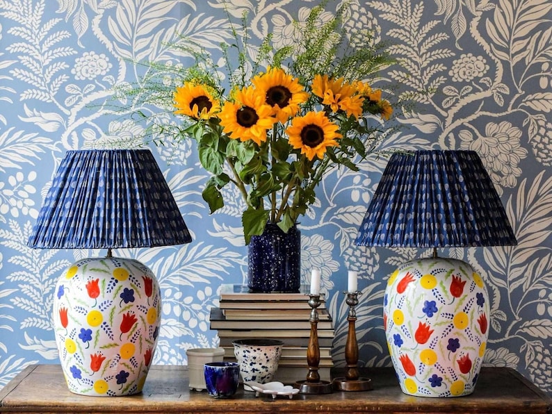 a table with two blue lamps, a vase of yellow sunflowers, and a stack of books. The table is set against a blue and white floral wallpaper.