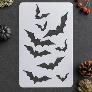 May include: A rectangular white stencil sheet featuring eight die-cut bat silhouettes of varying sizes. The bats are arranged in a scattered pattern across the surface, each displaying distinct wing shapes and pointed ears in a cutout design.