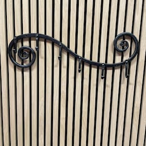 May include: A black metal wall-mounted coat rack with a decorative scroll design. The rack features six hooks and a curved, wave-like shape. The background is a light wood paneling with vertical black stripes.