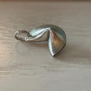 May include: A silver-colored fortune cookie charm with a small ring attached. The charm has a textured surface and is slightly open at one end. The charm is lying on a white wooden surface.