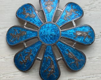 May include: A silver and blue enamel flower-shaped brooch with intricate designs. The center of the brooch is a circle with a blue enamel design. The petals of the flower are decorated with silver and blue enamel designs of figures.