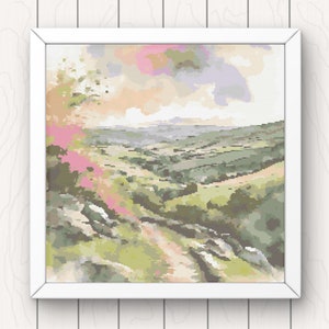 Puede incluir: Una bordado de punto de cruz enmarcado que representa un paisaje escénico con colinas onduladas, un camino sinuoso y un vibrante arbusto floreciente de color rosa en primer plano. El cielo es una suave mezcla de rosa, naranja y morado.