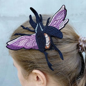 May include: A black and purple embroidered insect hair clip. The insect has detailed wings with purple and white accents. The hair accessory is attached to the hair of a person, with the back of the head and ear visible.