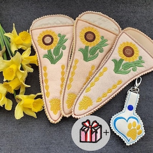 May include: Three beige fabric cases with embroidered sunflowers, green leaves, and yellow accents. A matching keychain with a heart and paw print design is included. A gift icon is also present. Yellow daffodils are in the background.
