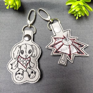 May include: Two embroidered keychains. One is a white dog with a red heart, the other is a white starburst design with red and black accents. Both have silver-tone clasps and white leather-like tags.