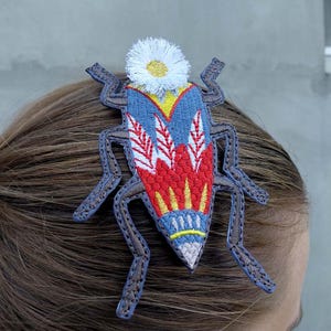 May include: A colorful embroidered insect-shaped hair clip. The clip features a blue, red, and yellow design with white accents and a white flower on top. The clip is attached to brown hair.