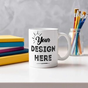 May include: A white ceramic mug with the text "Your DESIGN HERE" in black. The mug has a curved handle and sits on a white surface. Colorful books and paintbrushes in a jar are in the background.