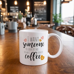 Puede incluir: Taza de caf&eacute; de cer&aacute;mica blanca con una inscripci&oacute;n negra que dice "Quiero que alguien me mire como yo miro al caf&eacute;". La taza tiene un dise&ntilde;o de coraz&oacute;n rosa y dorado.