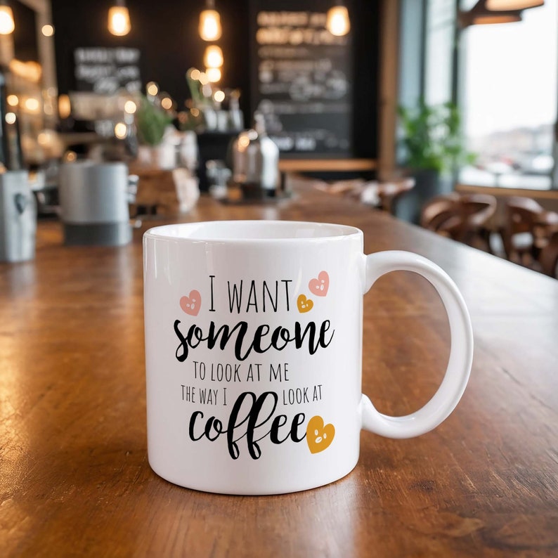 Puede incluir: Taza de caf&eacute; de cer&aacute;mica blanca con una inscripci&oacute;n negra que dice "Quiero que alguien me mire como yo miro al caf&eacute;". La taza tiene un dise&ntilde;o de coraz&oacute;n rosa y dorado.