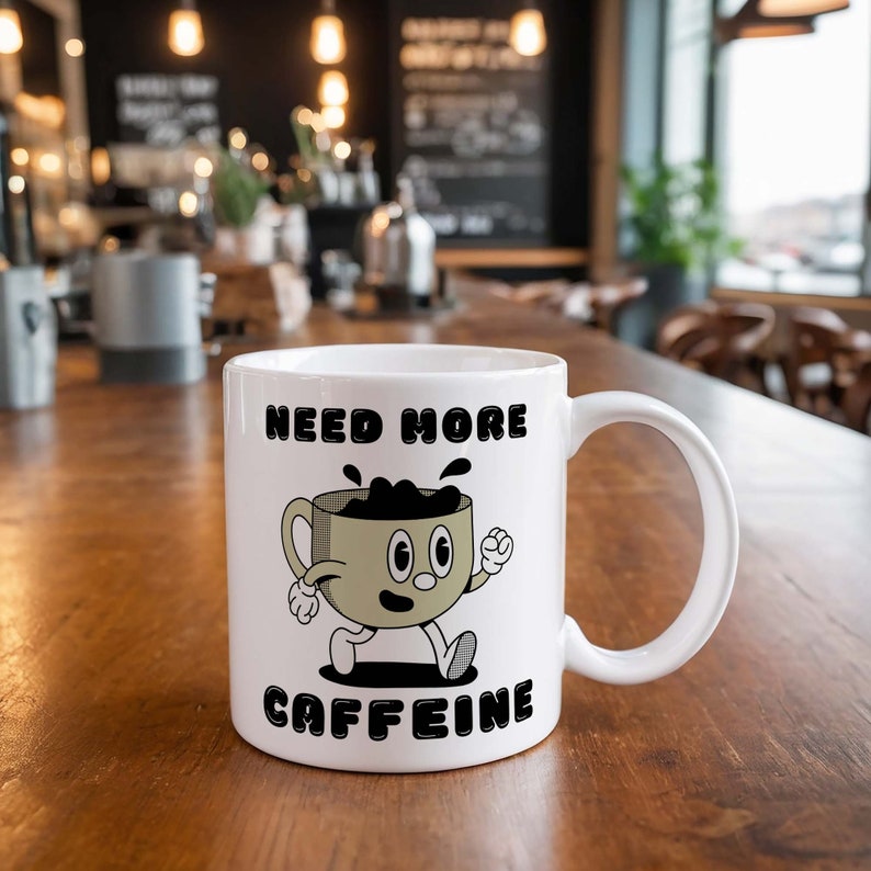 Puede incluir: Taza de caf&eacute; de cer&aacute;mica blanca con una ilustraci&oacute;n en blanco y negro de una taza de caf&eacute; con una cara y el texto "Need More Caffeine".