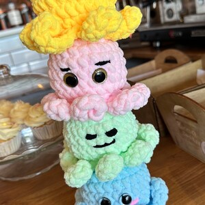 May include: A stack of four crocheted octopus plushies in yellow, pink, green, and blue. Each octopus has a different facial expression and black eyes.