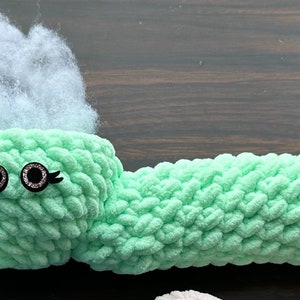 May include: A light green crocheted toy with black eyes and white eyelashes. The toy is shaped like a long, thin worm.