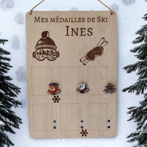 Peut inclure: Une plaque en bois avec le texte "Mes médailles de Ski Ines" gravé dessus. La plaque a un design de lunettes de ski et de bâton de ski en haut et un espace pour les médailles avec un flocon de neige en bas.