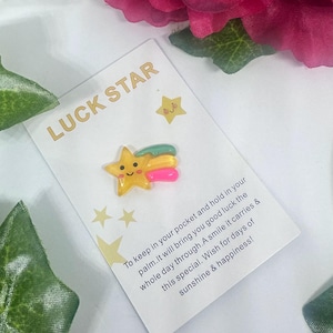 May include: A white card with gold lettering reading "LUCK STAR". A yellow star charm with a pink and green trail is attached. The card has a message about good luck and happiness. The background includes flowers.