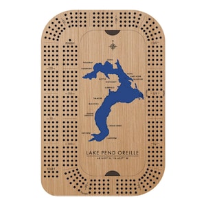May include: Wooden cribbage board featuring a blue map of Lake Pend Oreille with place names. The board has a light wood grain finish, a compass rose, and scoring holes. Text includes "Lake Pend Oreille" and coordinates.