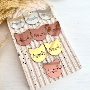 Puede incluir: Un conjunto de siete pendientes de acrílico con forma de gato con la palabra "Kajusky" escrita en cada uno. Los pendientes son de varios colores, incluyendo plata, oro y oro rosa.