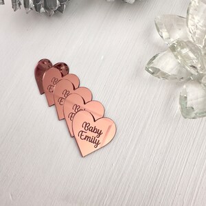 May include: Five rose gold heart-shaped acrylic place cards with the text "Baby Emily" on one of the cards.