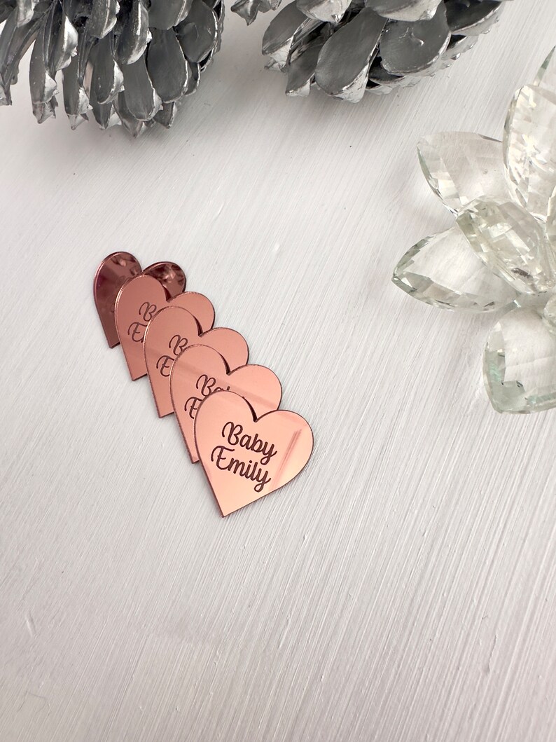 May include: Five rose gold heart-shaped acrylic place cards with the text "Baby Emily" on one of the cards.