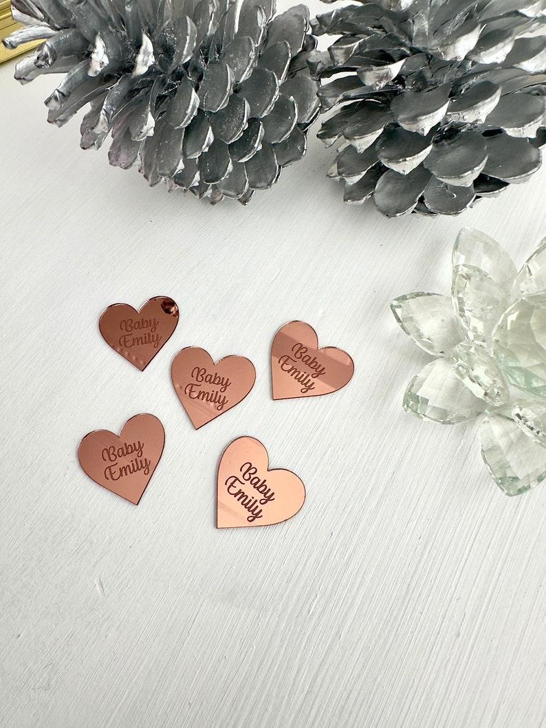 May include: Five rose gold heart-shaped acrylic ornaments with the text "Baby Emily" engraved on each.