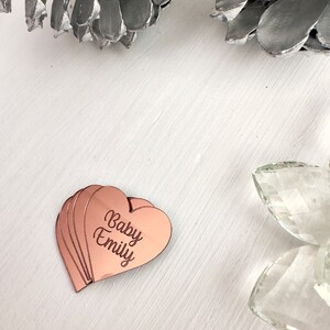 May include: A set of rose gold heart-shaped acrylic plaques with the text "Baby Emily" engraved on them.