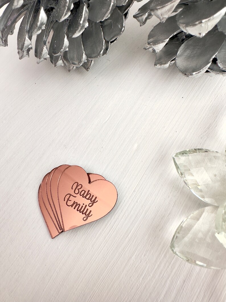 May include: A set of rose gold heart-shaped acrylic plaques with the text "Baby Emily" engraved on them.