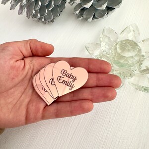 May include: A set of five rose gold heart-shaped mirror tiles with the text "Baby Emily" engraved on them.