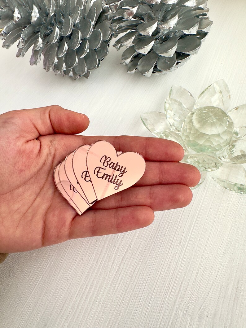 May include: A set of five rose gold heart-shaped mirror tiles with the text "Baby Emily" engraved on them.