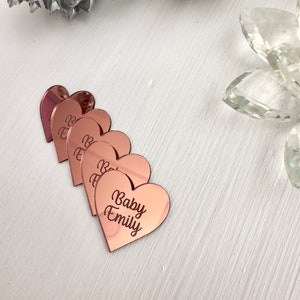 May include: Five rose gold heart-shaped acrylic place cards with the text "Baby Emily" engraved on the bottom card.