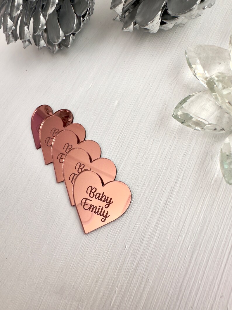 May include: Five rose gold heart-shaped acrylic place cards with the text "Baby Emily" engraved on the bottom card.