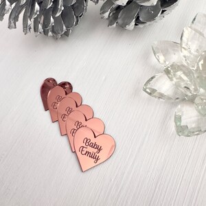 May include: Five rose gold heart-shaped acrylic place cards with the text "Baby Emily" on the largest heart.