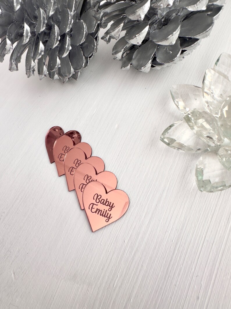May include: Five rose gold heart-shaped acrylic place cards with the text "Baby Emily" on the largest heart.