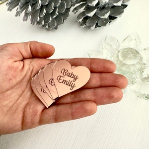 May include: A stack of rose gold heart-shaped acrylic place cards with the text "Baby Emily" engraved on them.
